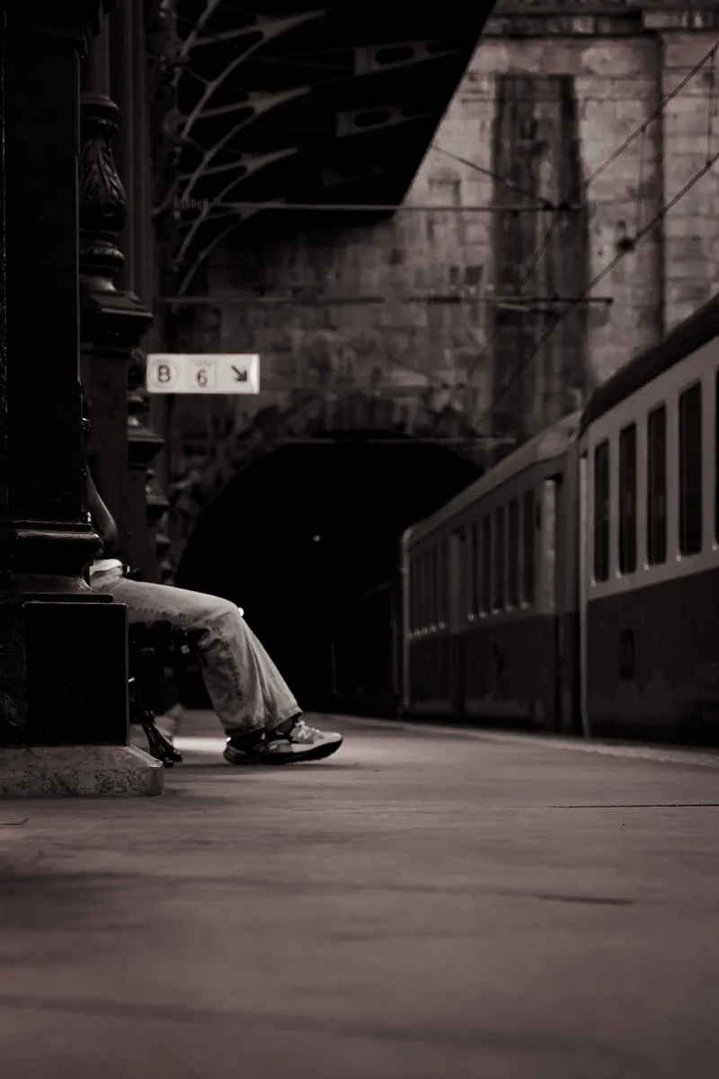 Black and white image of train platform with solitary foot resting on bench (Mustvalge pilt rongijaamast, pingil lebav jalg – ootusmoment)