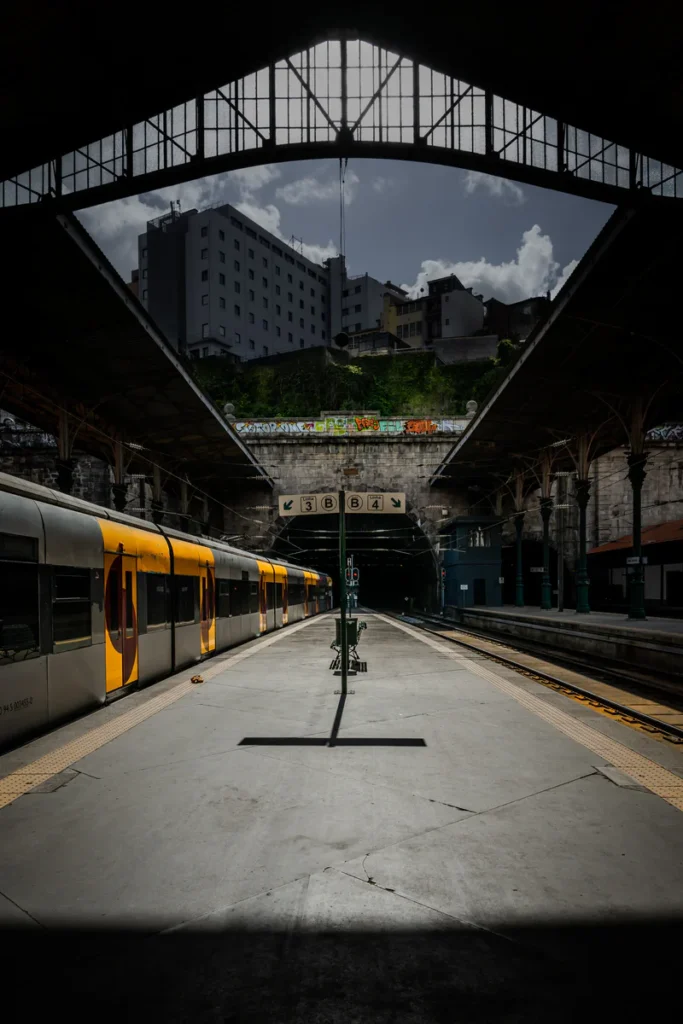 A yellow train stands beneath a large metal roof beside a sunlit platform.