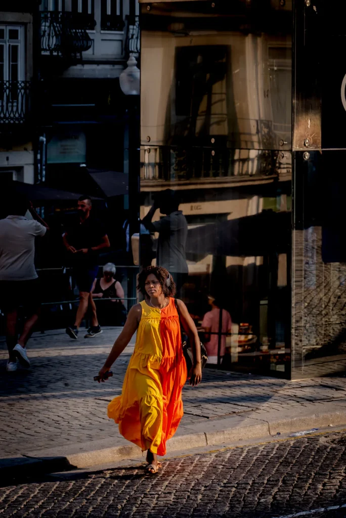 A woman in a yellow dress steps into a patch of sunlight on a cobbled street.