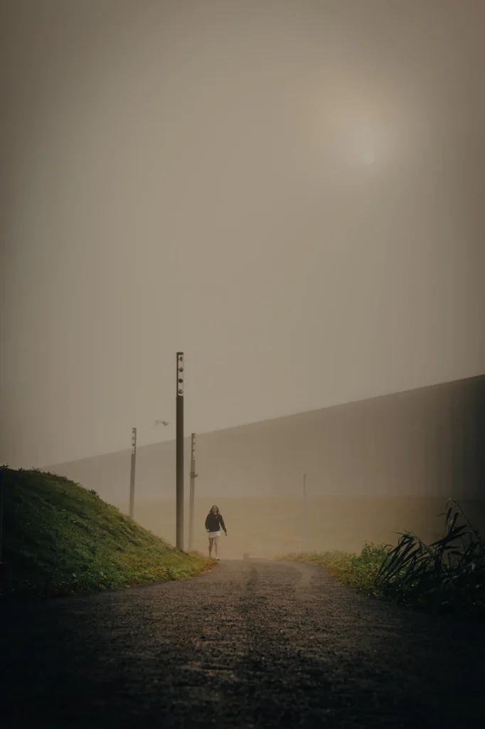 One person walks along a misty gravel path in front of a building with a long glass facade.