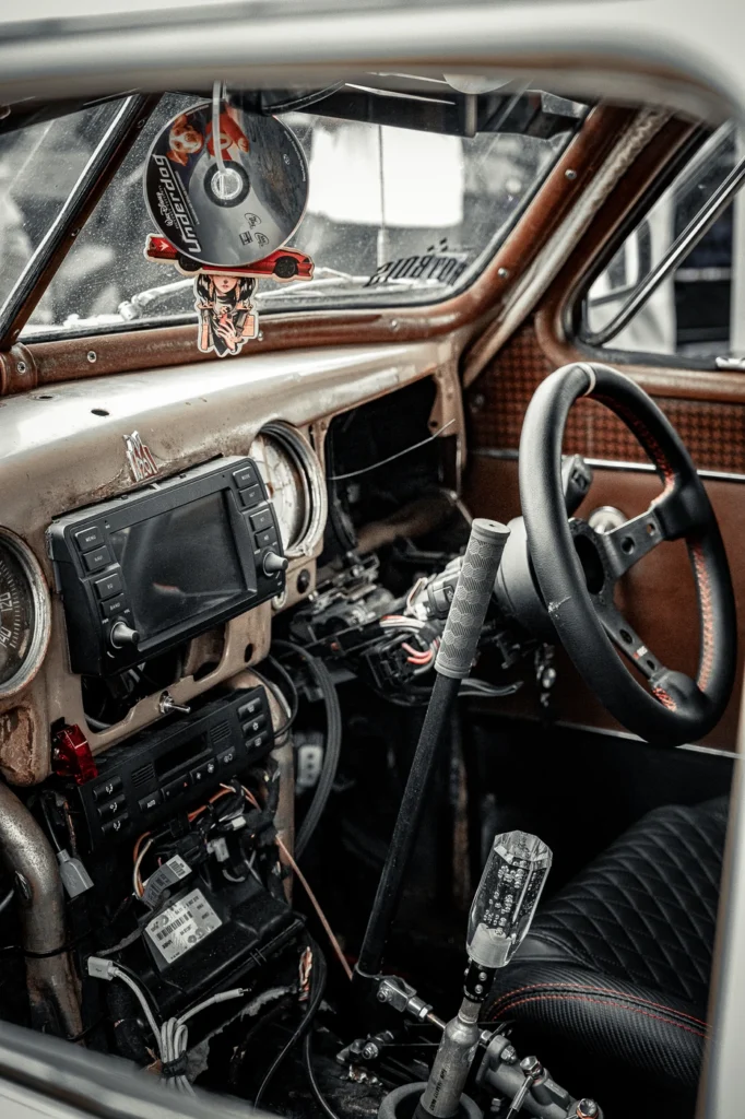 Worn dashboard of a vintage car with an added screen, sports steering wheel and long gear lever.