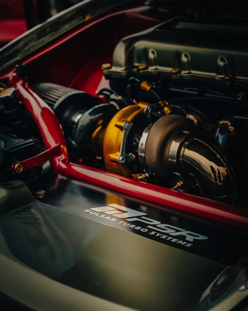 Close-up of a turbocharger, metal pipework and a red brace in an engine bay.