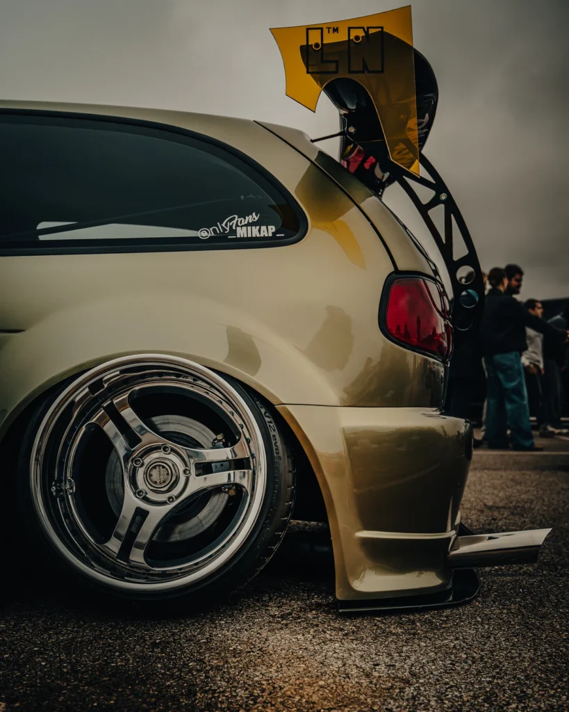 Low rear quarter of a gold car with a polished wheel and tall rear wing.