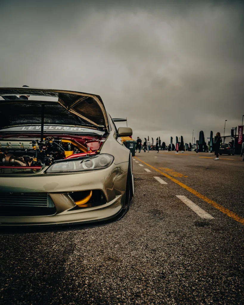 Open bonnet and engine bay of a gold modified car, with festival visitors in the background.