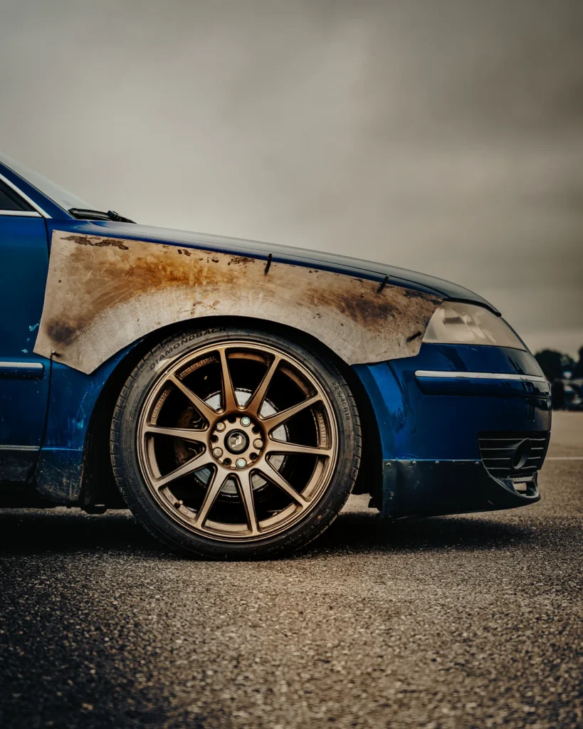 Front wheel of a blue car with a bronze-coloured rim and rusty metal wing.