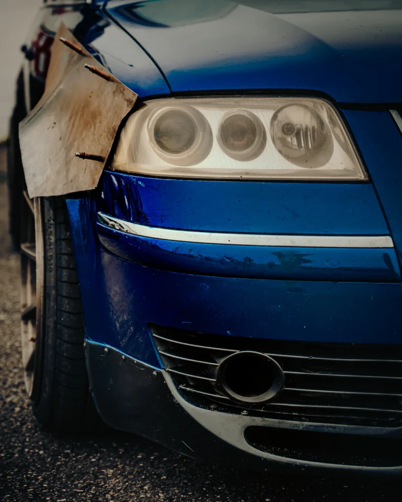 Headlight and front bumper of a blue car beside a metal wing held by visible fasteners.