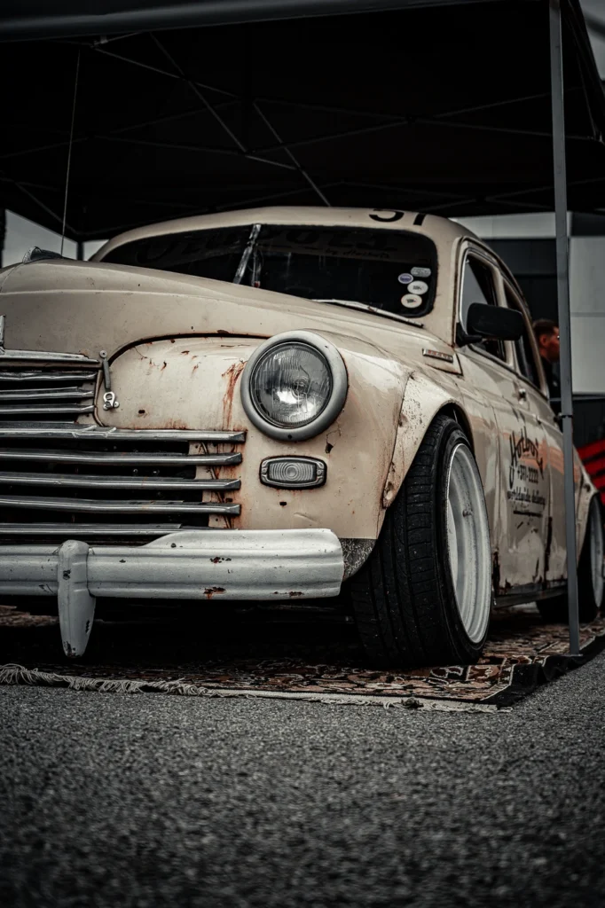 A pale vintage car on a patterned rug, photographed close to the ground.