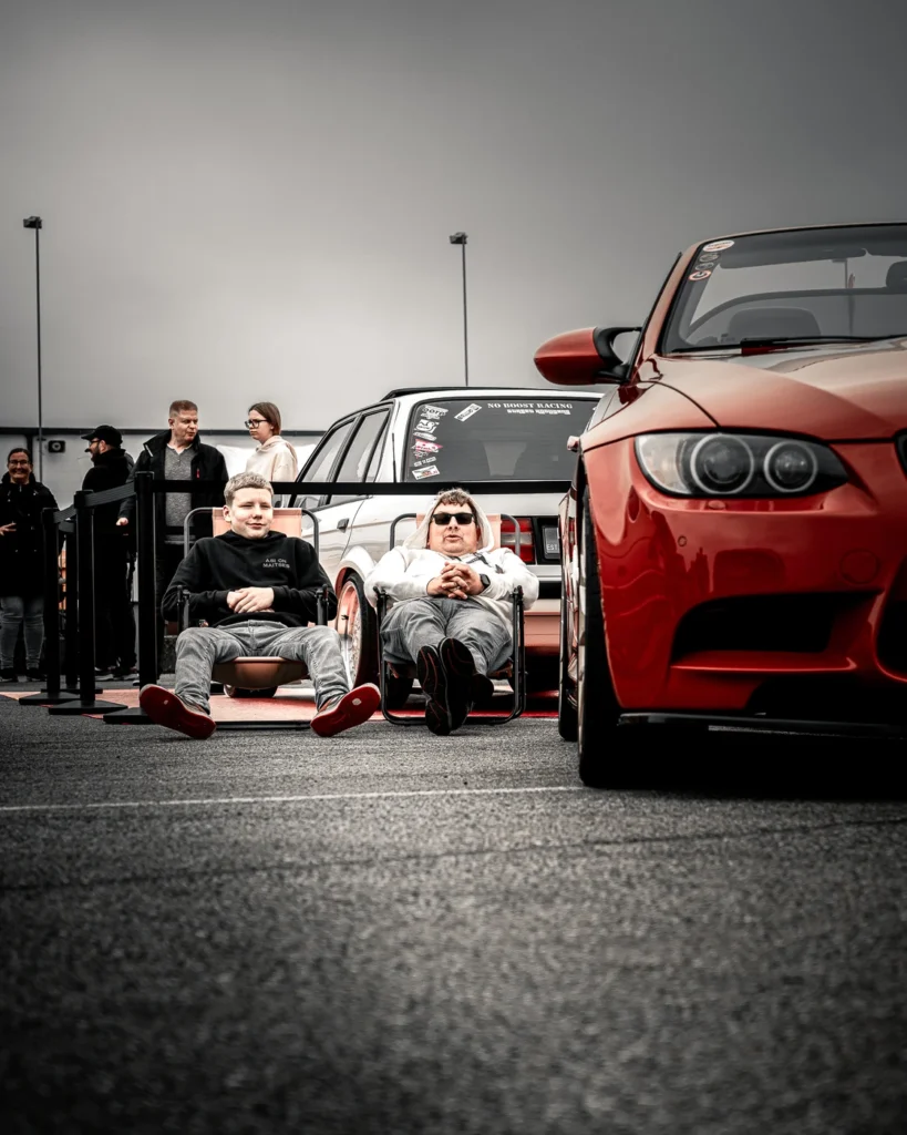 Two visitors sitting on low chairs beside a red BMW.