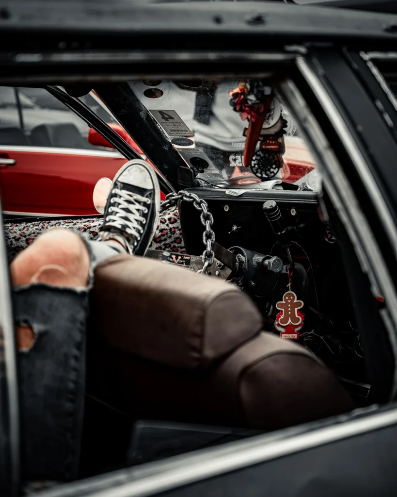A visitor resting on a brown car seat with a foot in a trainer towards the dashboard decorated with chains.