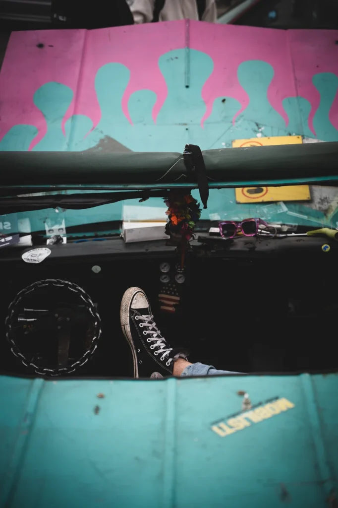 View into a turquoise car with a foot in a trainer inside and a raised bonnet painted with flames behind it.