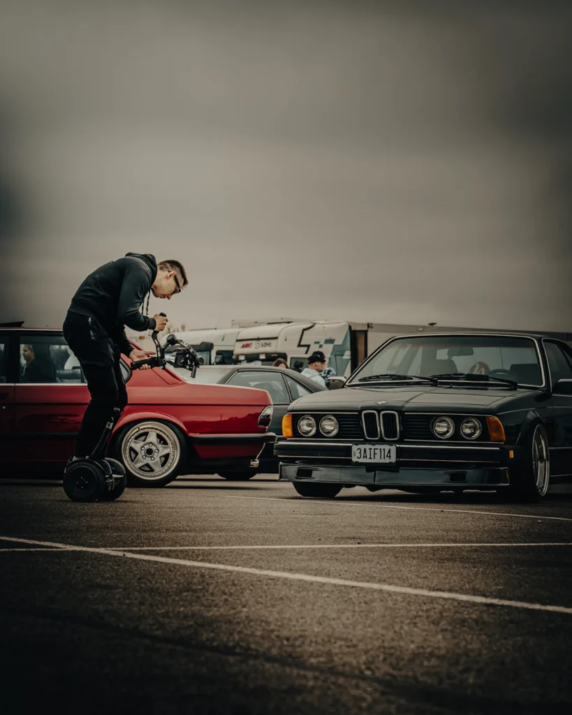 A person filming from a self-balancing scooter in front of a black BMW, beside a red convertible.