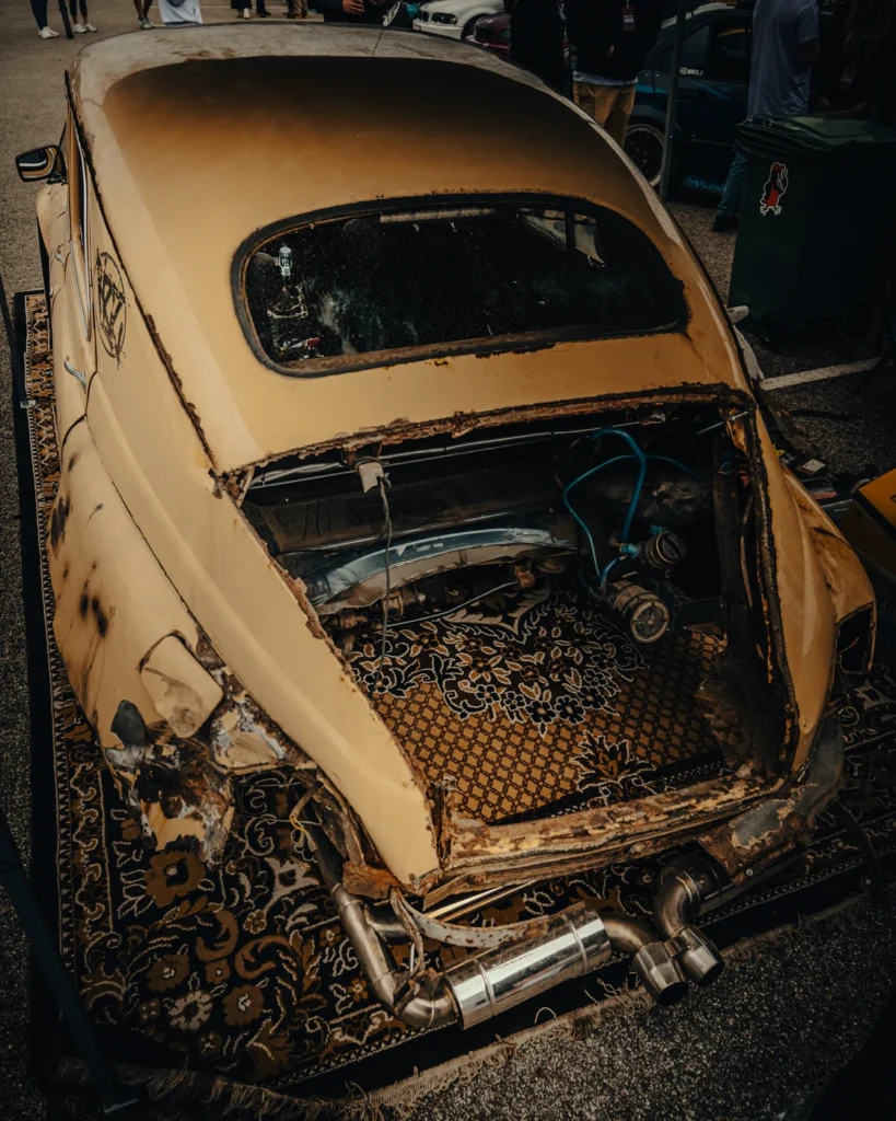 Open rear compartment of a yellowish vintage car with exposed components and a patterned rug over the rear edge.