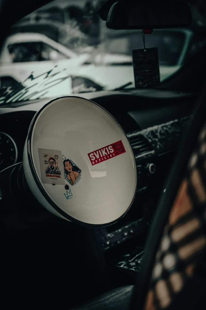 White bowl covered in stickers in place of a car steering wheel, with the dashboard and rear-view mirror behind it.