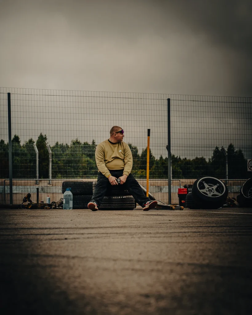 Visitor in a pale sweatshirt sitting on stacked tyres by a wire fence, beside wheels and tools.