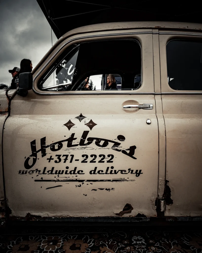 Door of a pale vintage car with rust patches, Hotbois lettering and a black side mirror.