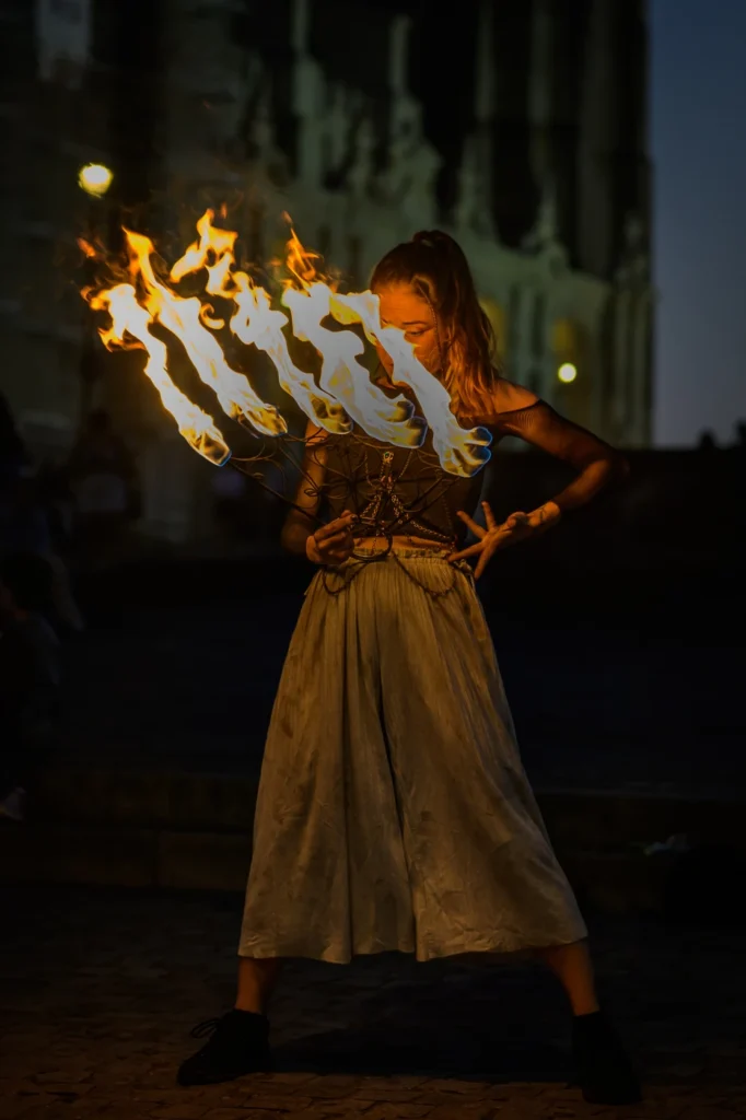 Natalia holds burning fans in front of her chest, with a dim building facade behind her.
