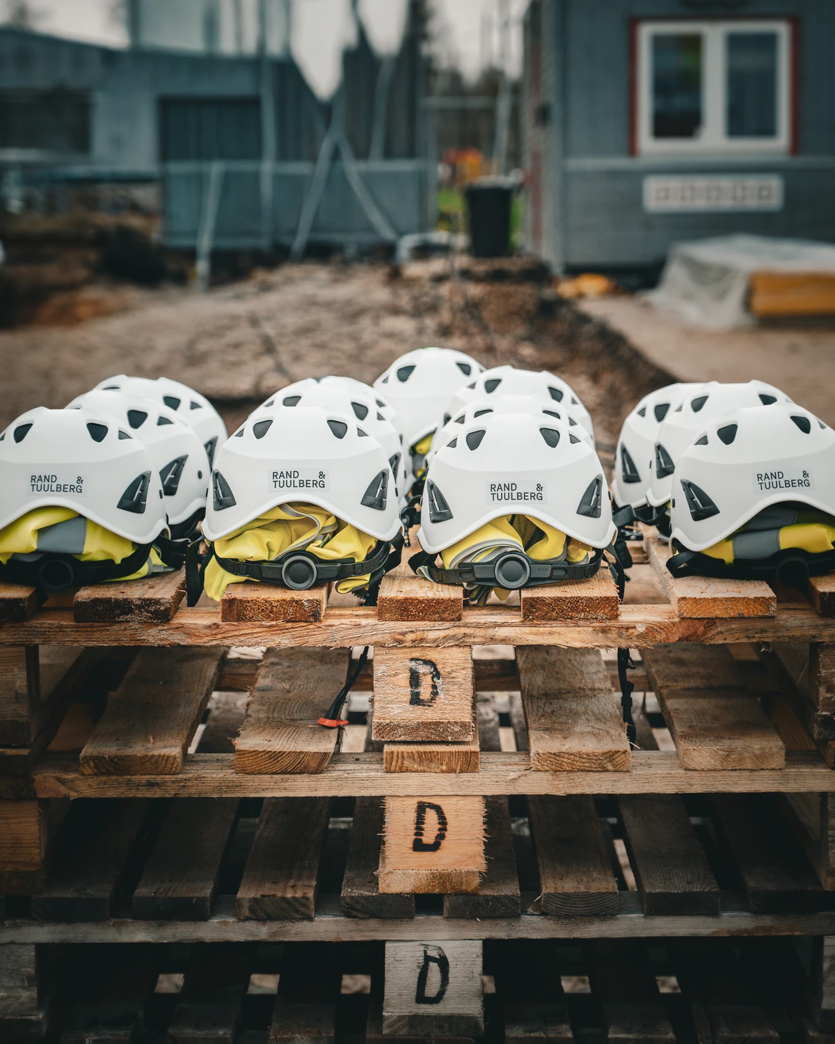 White Rand & Tuulberg hard hats and yellow safety vests on wooden pallets in front of a construction site.