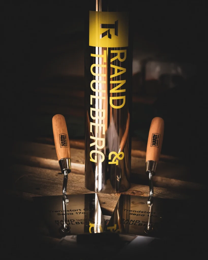 A polished cornerstone capsule bearing the Rand & Tuulberg name, flanked by two wooden-handled trowels.
