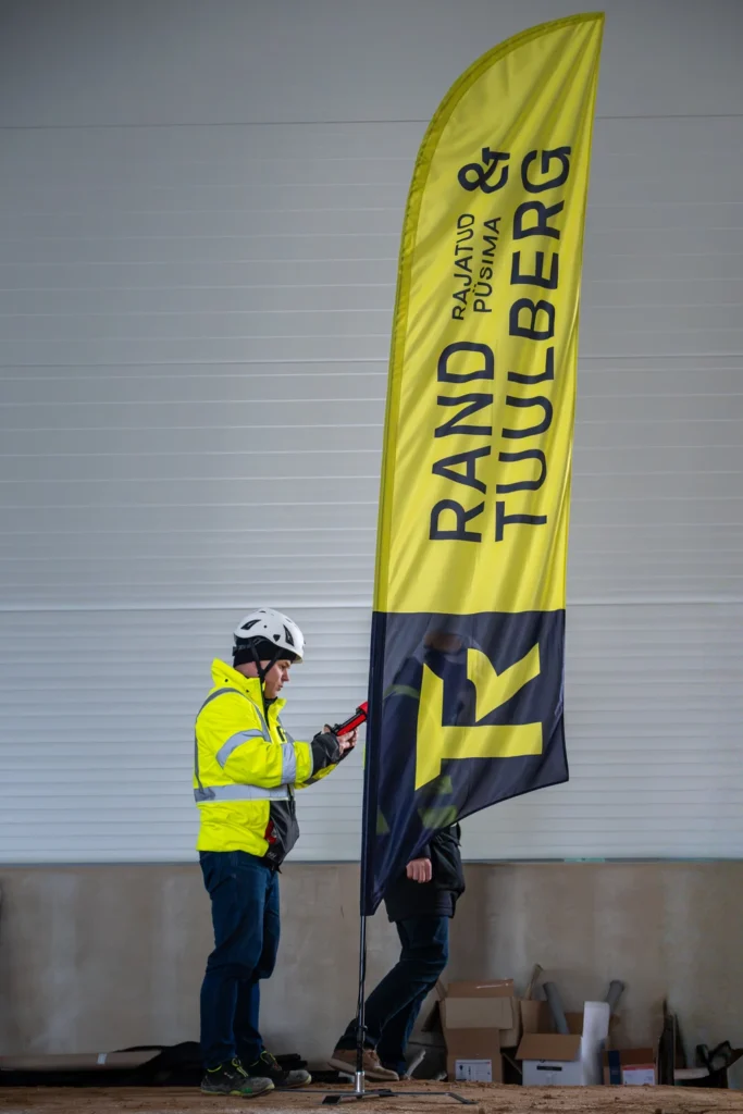 Two people beside a tall yellow Rand & Tuulberg flag inside a building under construction.