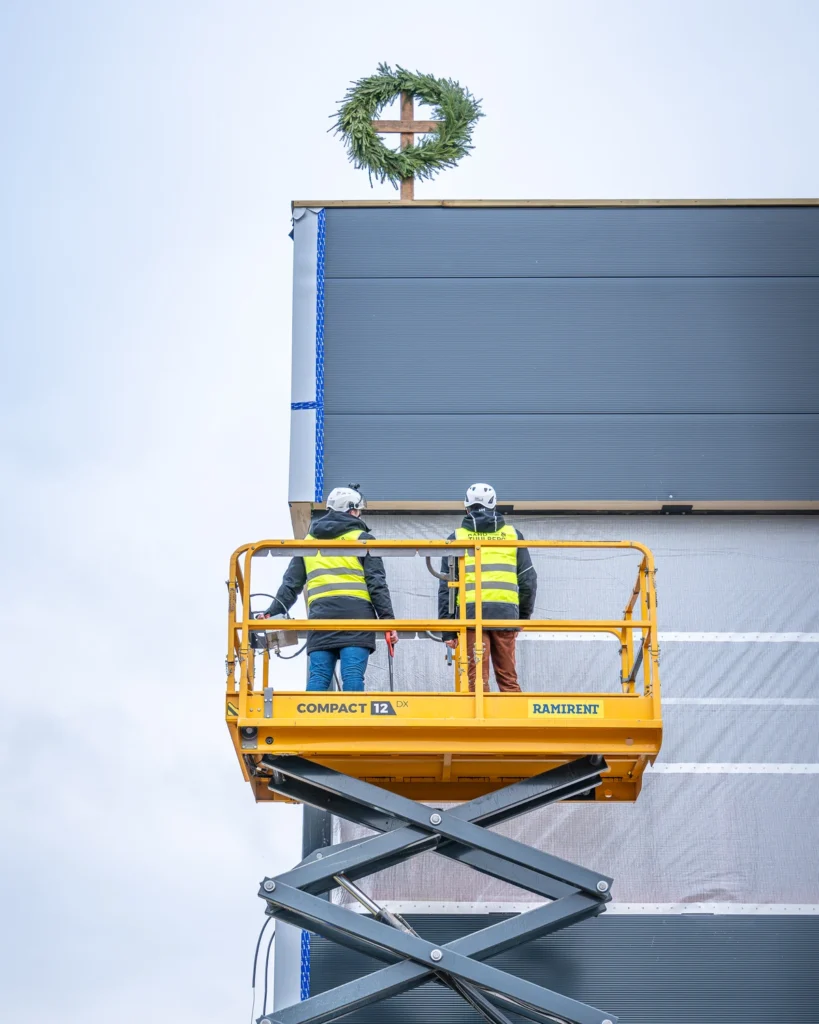 Two people in hard hats on a scissor lift, with a green topping-out wreath on the roof above them.