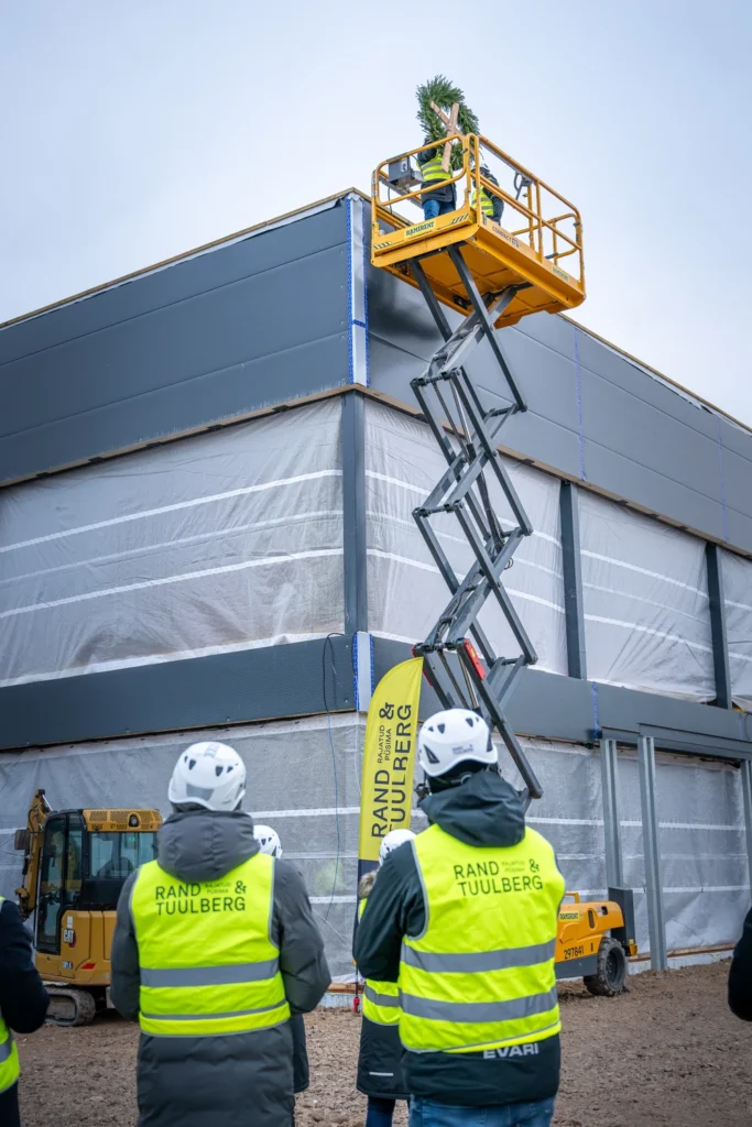 A scissor lift raised beside the corner of a building, watched by two people on the ground.
