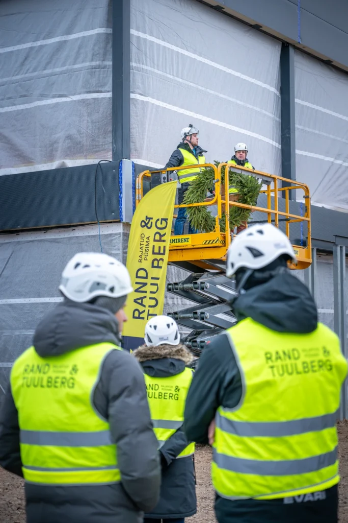 A topping-out wreath and two people on a scissor lift, with spectators in hard hats in the foreground.