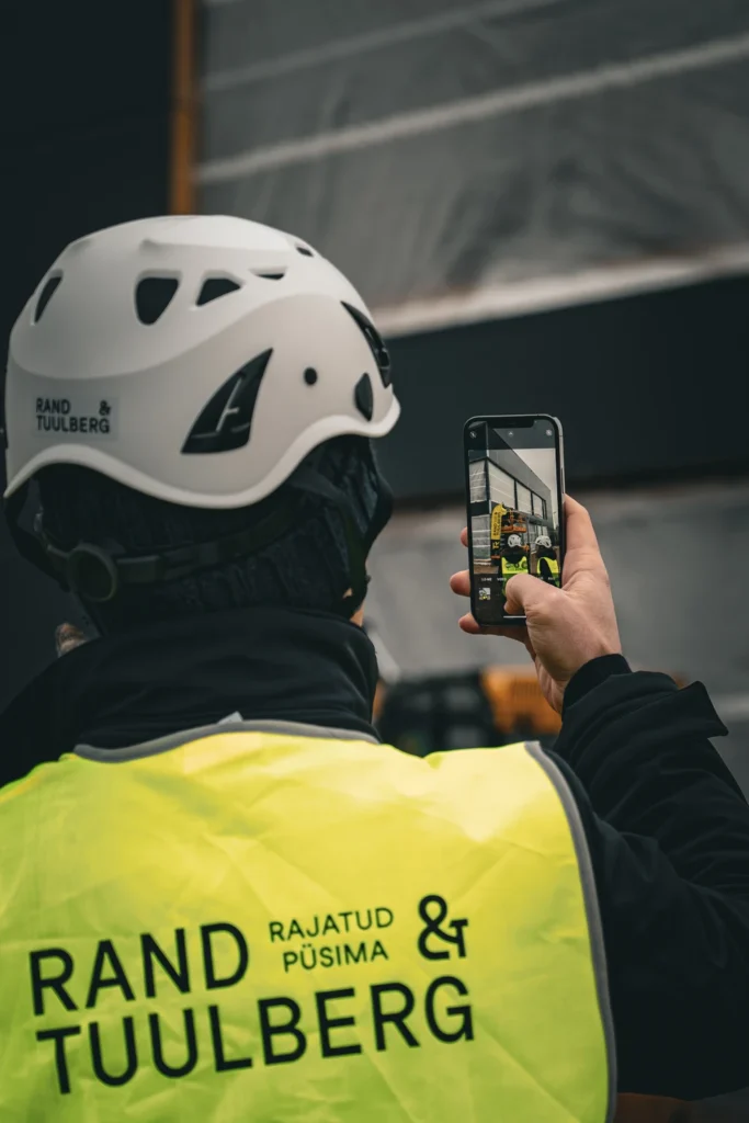 A person in a Rand & Tuulberg hard hat and safety vest records activity on the construction site with a phone.