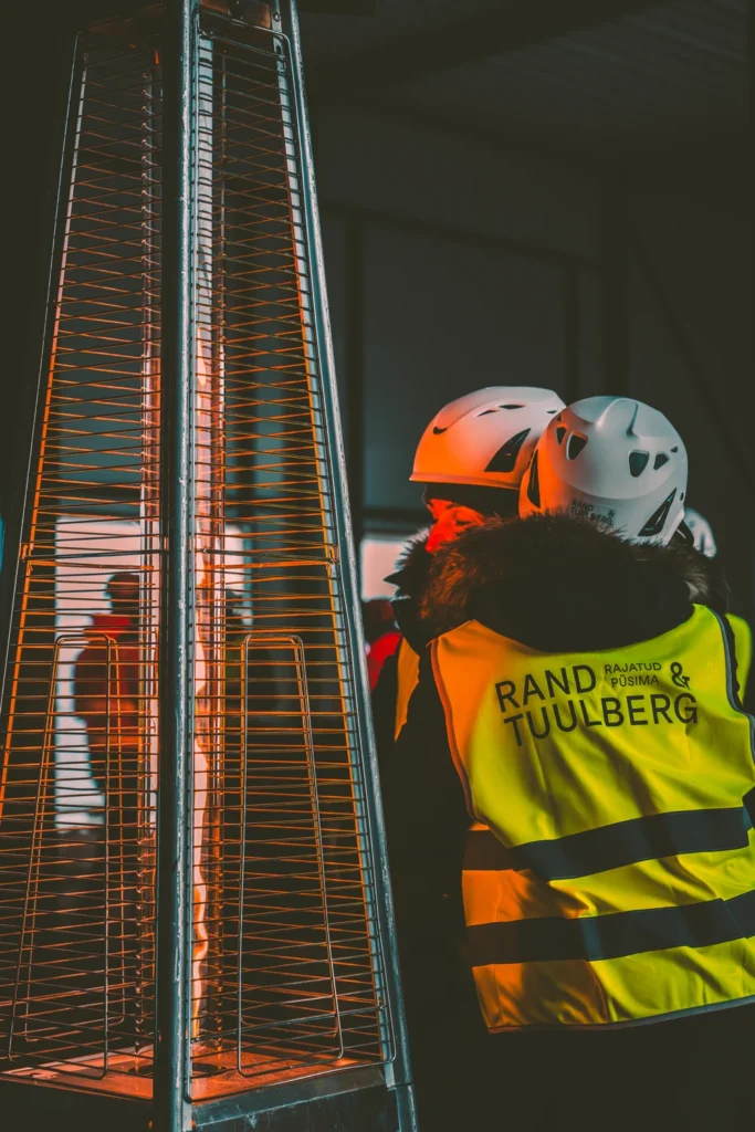 Two people in hard hats and safety vests stand beside a glowing gas heater.