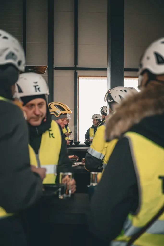 People in hard hats and yellow safety vests gather around tables inside a building under construction.