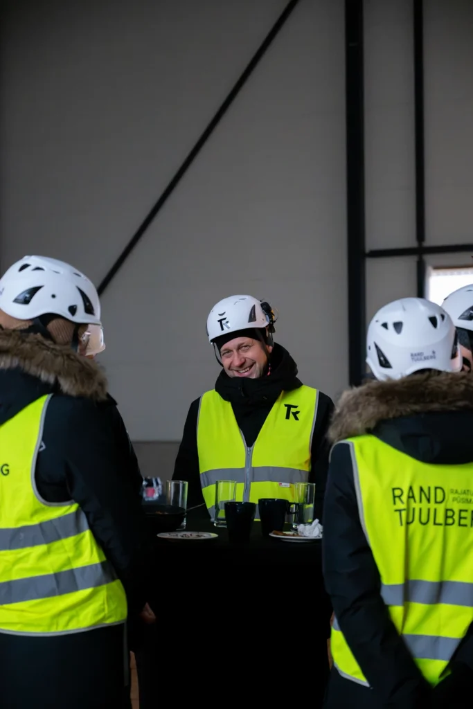 A smiling man in a hard hat talks with people in safety vests gathered around a table.