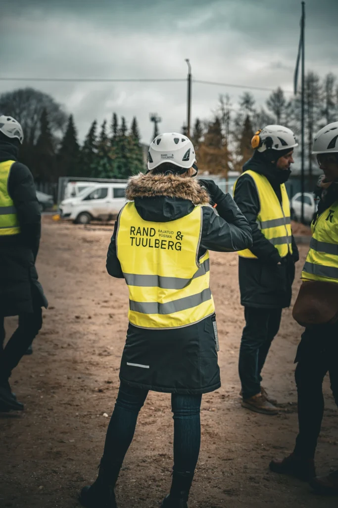 A person in a yellow Rand & Tuulberg vest, seen from behind among other attendees on the construction site.