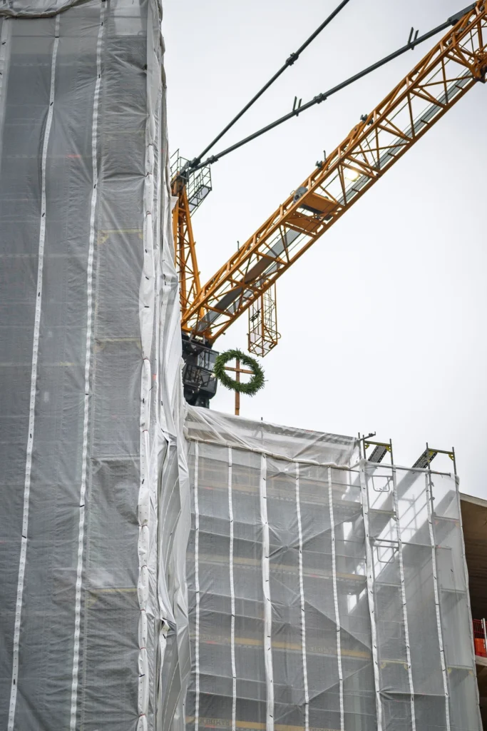 A building wrapped in scaffold sheeting, with a tower crane and a topping-out wreath above it.