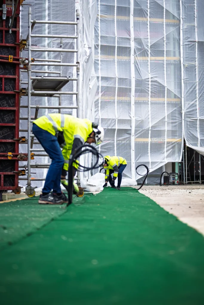 Two people in high-visibility jackets work beside a green carpeted walkway, one holding a coil of black cable.