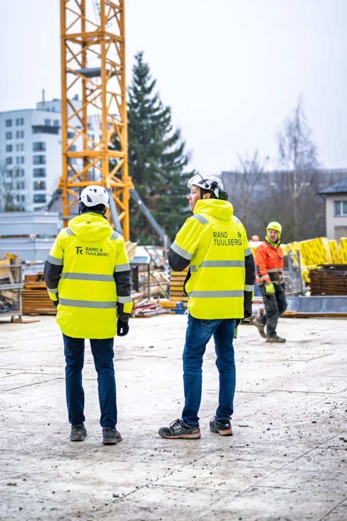 Two people in yellow Rand & Tuulberg jackets stand near the base of a tower crane on the construction site.