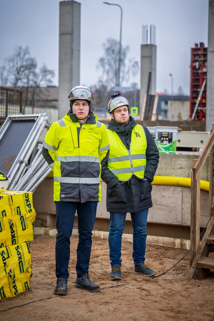 Two men in hard hats and high-visibility clothing stand among the concrete columns of a building under construction.
