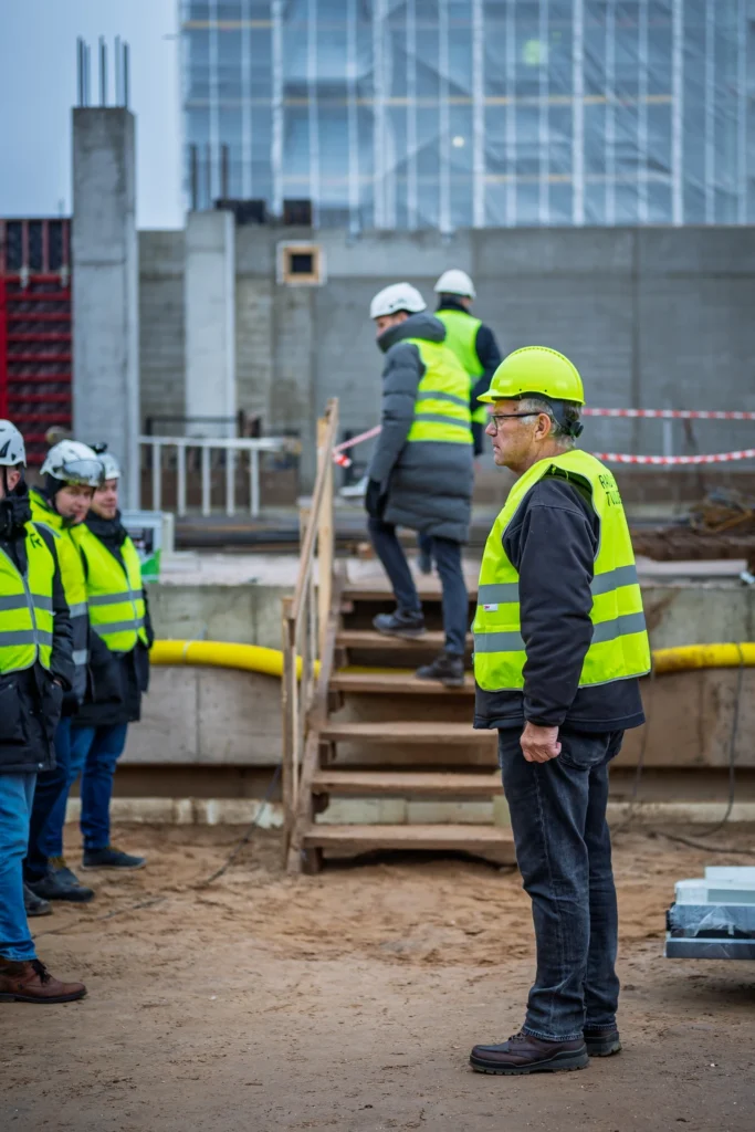 A man in a green hard hat stands beside temporary wooden steps as other attendees move behind him.