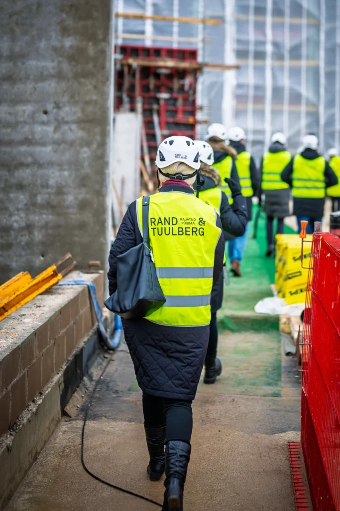 Attendees in safety vests and hard hats walk in single file along a narrow construction-site walkway.
