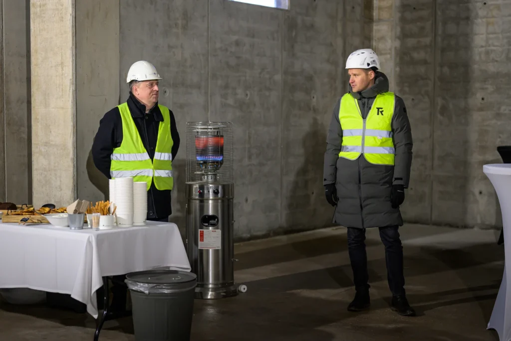 Two men in hard hats stand beside a refreshment table and a gas heater in a room with concrete walls.