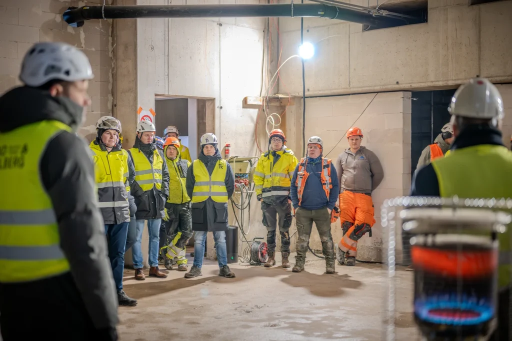People in high-visibility clothing and hard hats of different colours gather in a lit room under construction.