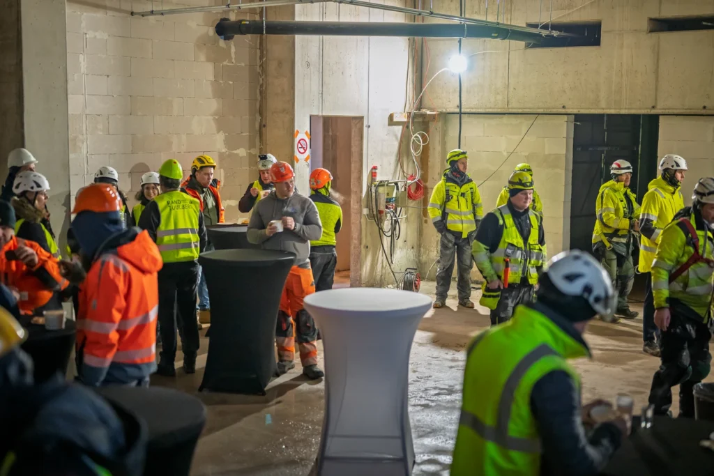 Topping-out celebration attendees chat and drink from cups around tall tables in an unfinished room.