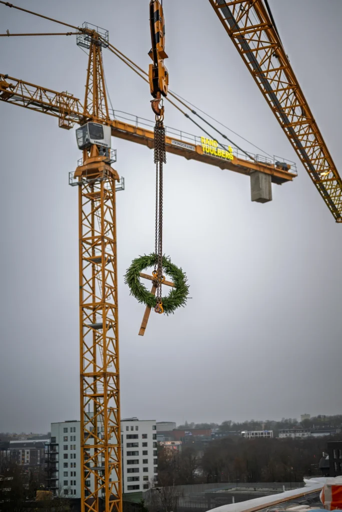 A green topping-out wreath hangs from a tower crane hook high above the construction site.