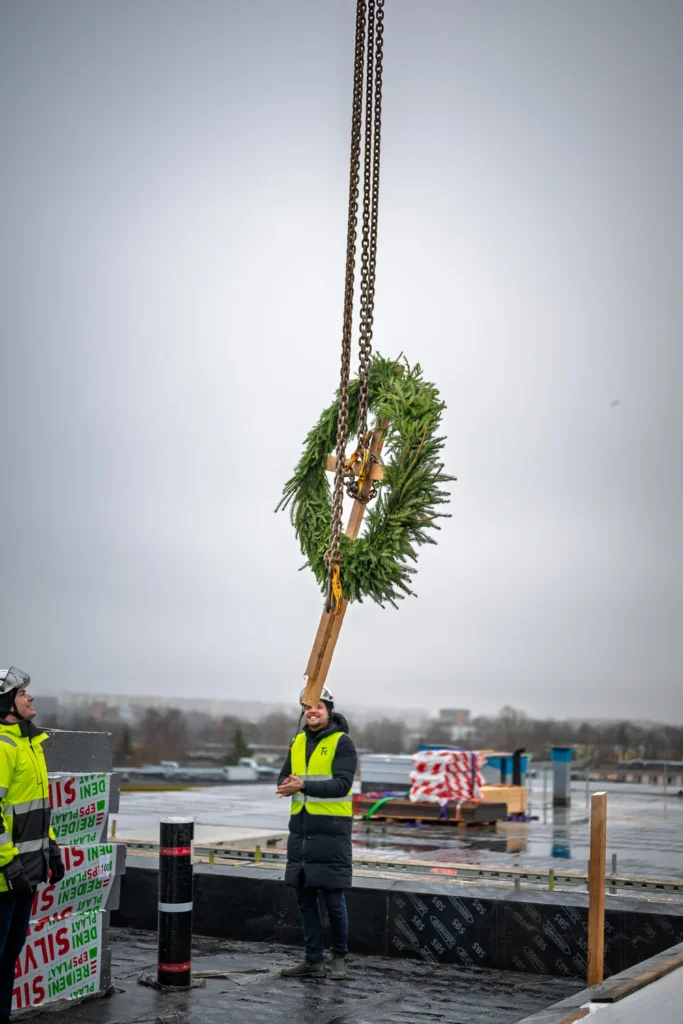 A topping-out wreath and wooden pole suspended from crane chains above a person standing on the roof.