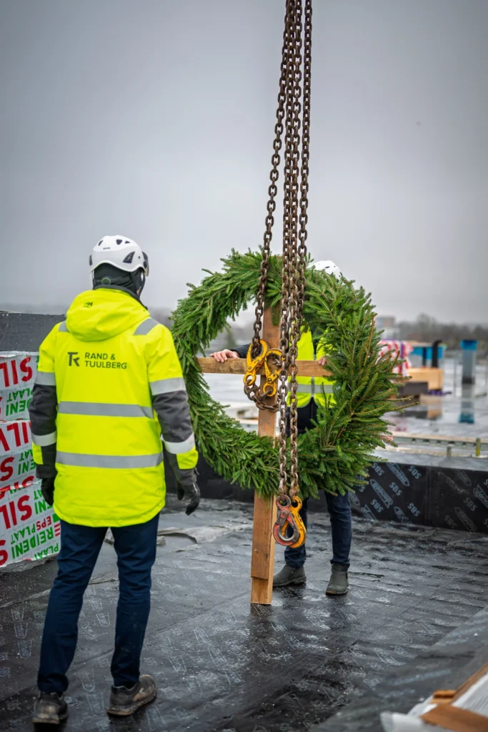 Crane chains and hooks hang in front of a topping-out wreath, with two people in high-visibility clothing on the roof.