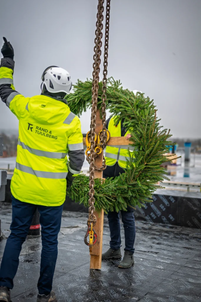 A person in a yellow high-visibility jacket points upward beside crane chains and a topping-out wreath.