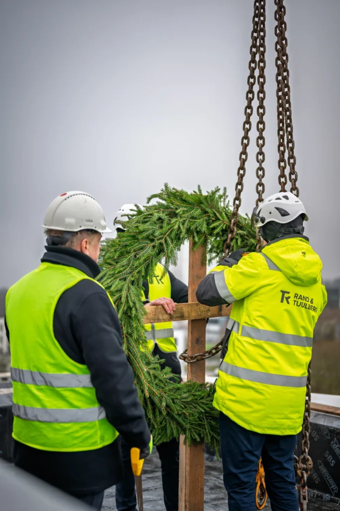 People in hard hats stand around a topping-out wreath as one handles the crane chains beside it.