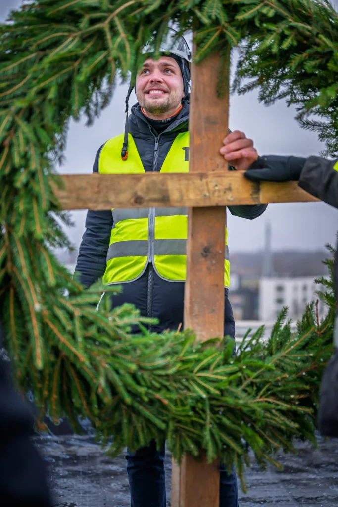 A smiling man in a hard hat holds a wooden pole and looks up through a green topping-out wreath.