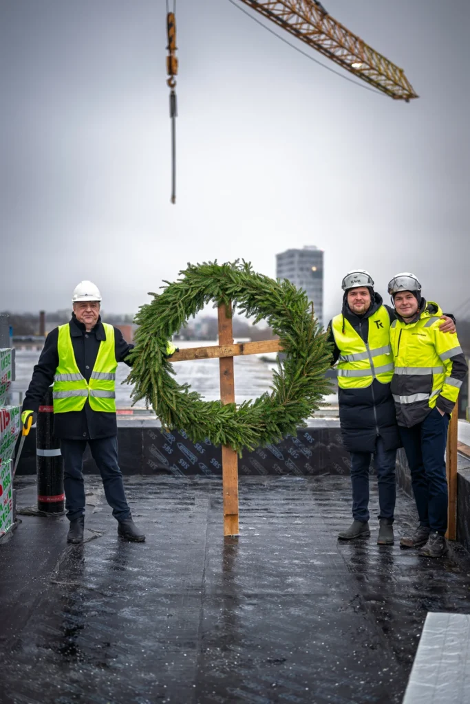 Three men in hard hats pose on either side of a large topping-out wreath on a rooftop, with a crane jib above.