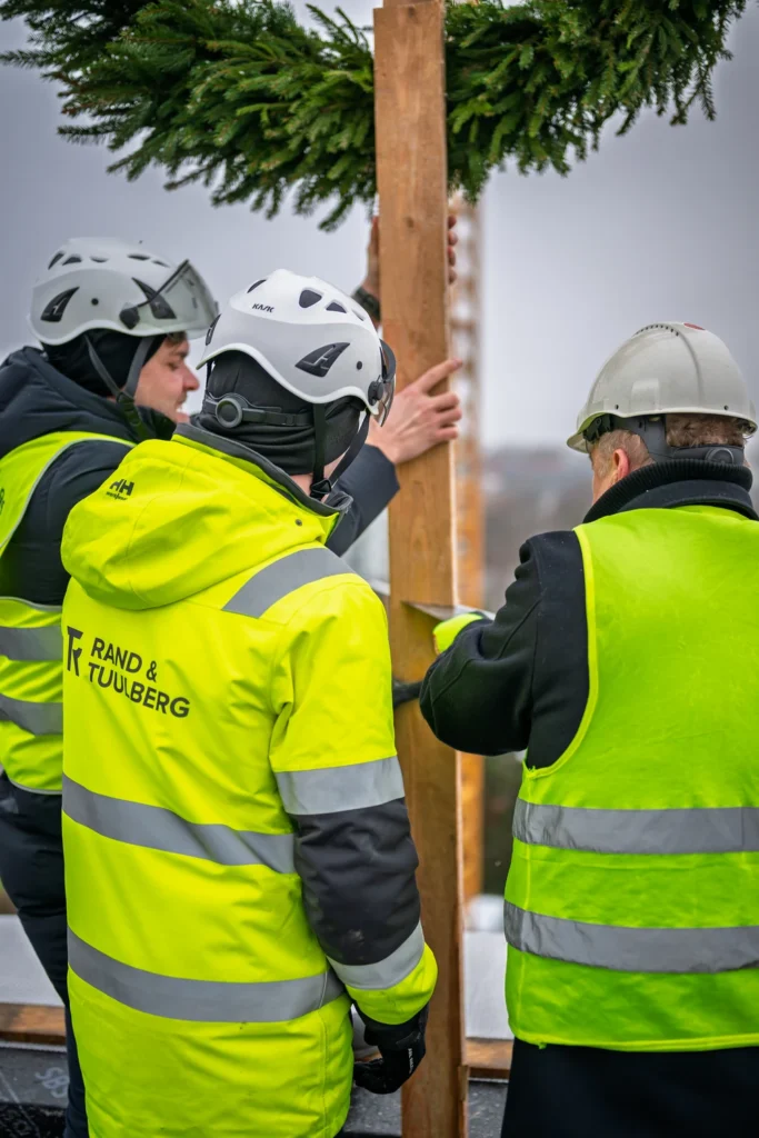 Three people in hard hats hold the pole supporting a green topping-out wreath, seen from behind.