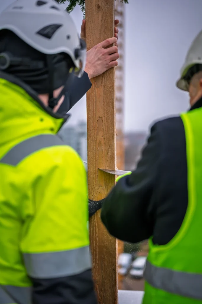 Close-up of hands, a saw and the wooden wreath pole between two people in hard hats.