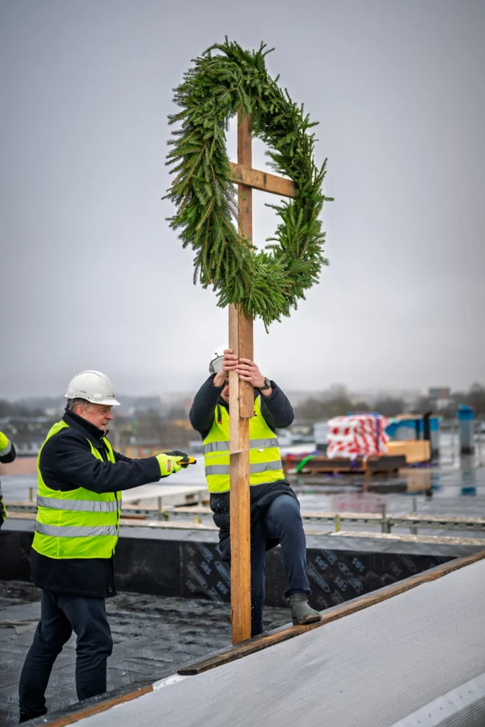 One man saws through a topping-out wreath pole while another holds it upright near the roof edge.